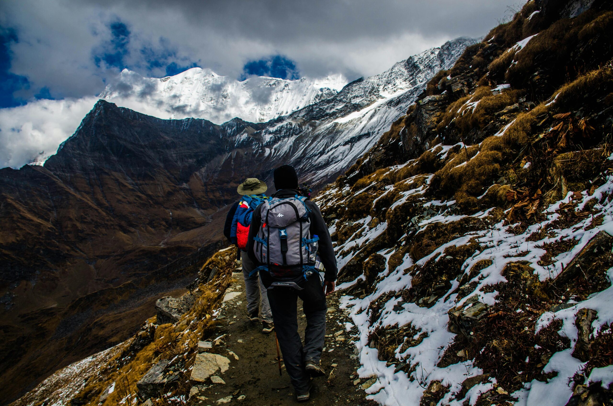 Home Two hikers navigating a snowy trail in rugged mountainous terrain under dramatic skies.